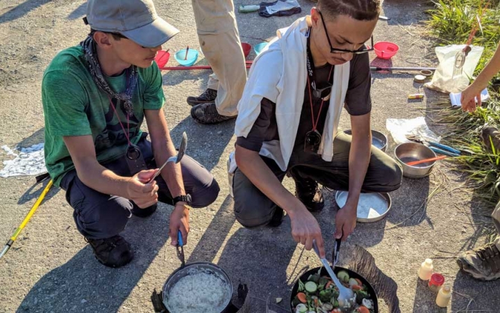 Two students prepare food using camping stoves.